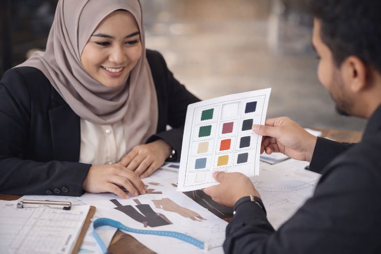 Perbincangan spesifikasi pakaian antara pelanggan wanita Melayu dan wakil Eraine dalam suasana meeting profesional menggunakan sampel warna dan dokumen rekaan.